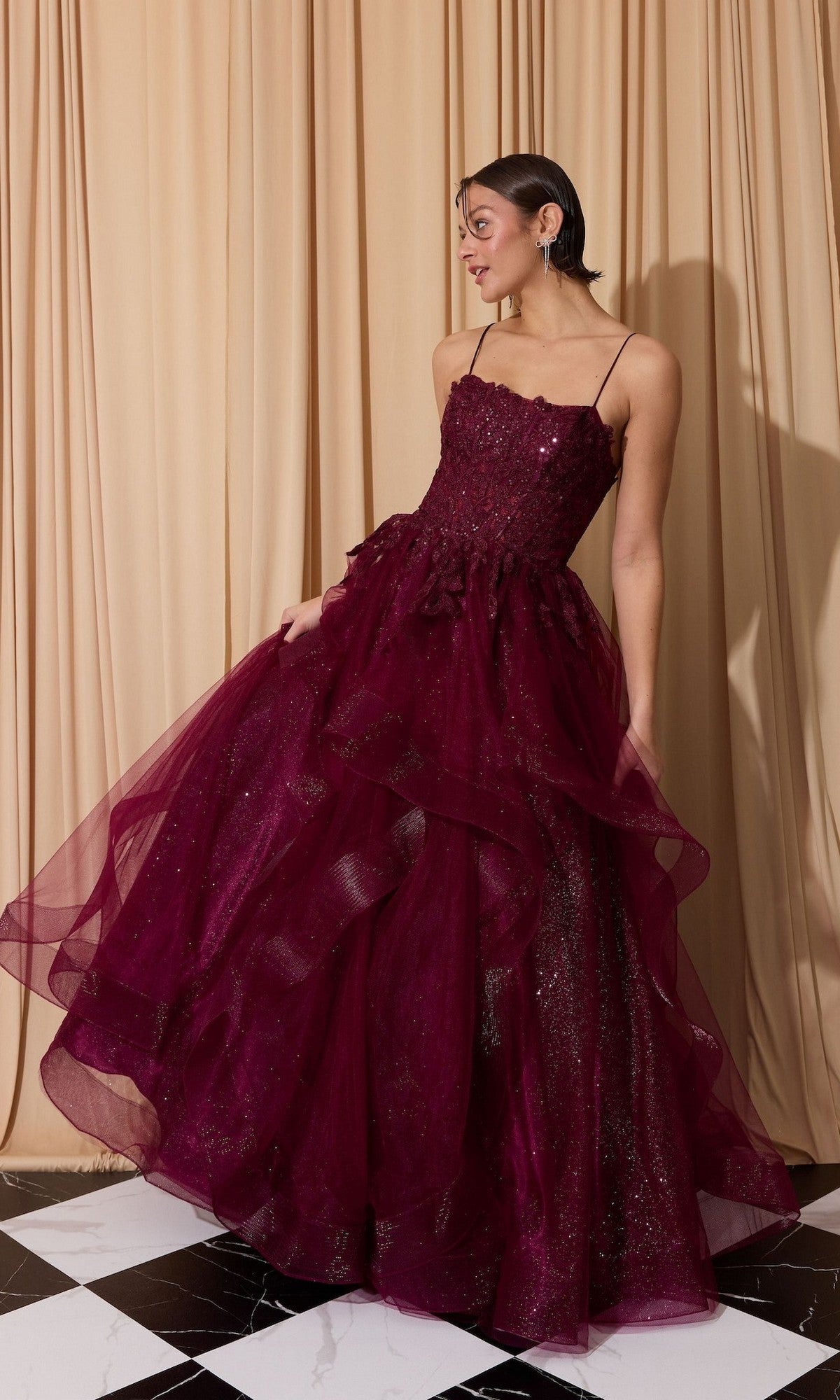 A woman poses on a black-and-white checkered floor in front of beige curtains, wearing the Long Prom Dress: Eureka EK167 in Berry—a glitter tulle gown with a floral lace embroidered corset and layered tulle A-line skirt.